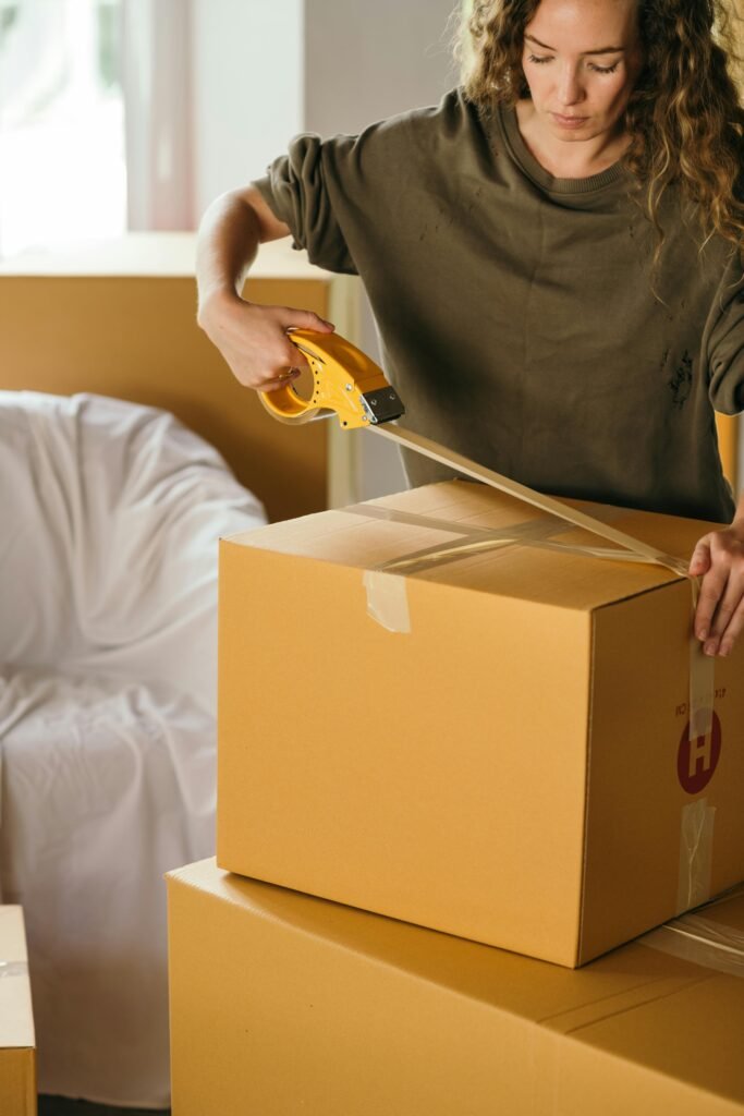 Crop female wearing casual clothes standing while packing carton boxes with scotch tape in modern apartment during preparing for relocation